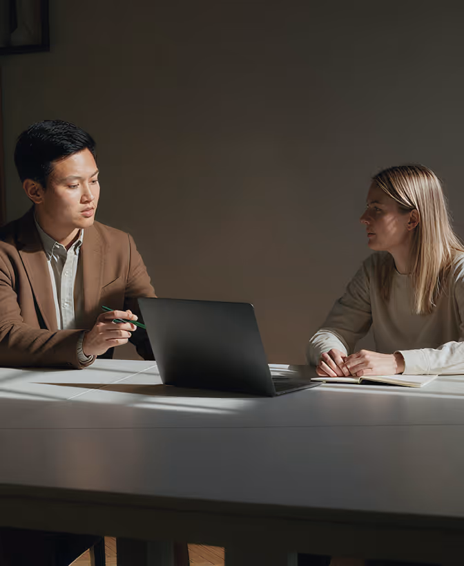 Man and woman sitting across a table with a laptop, engaged in a serious conversation in a dim room.