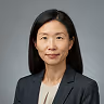 Portrait of a woman with straight black hair wearing a black blazer and necklace against gray background.